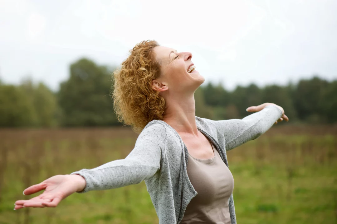 Žena s rozpaženýma rukama se směje v přírodě, vyjadřuje radost a svobodu na zelené louce pod širým nebem.