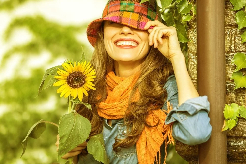 Žena v klobouku s úsměvem drží slunečnici, oblečená v džínové košili a oranžové šále, pozadí tvoří zeleň.