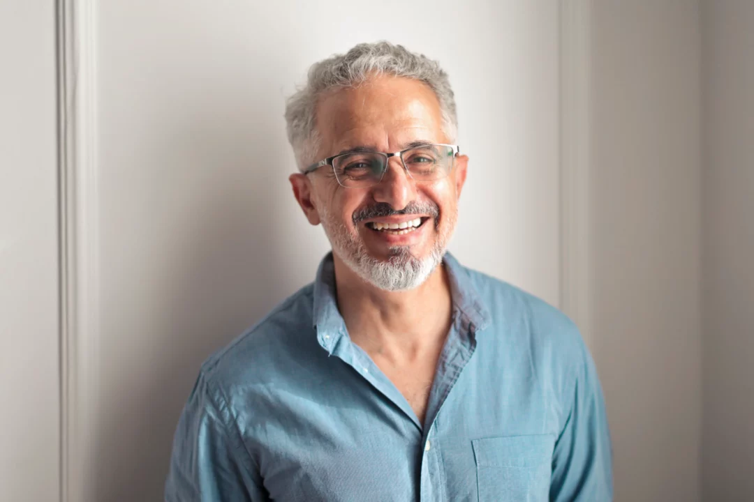 Usměvavý muž s brýlemi a šedivými vlasy v modré košili stojí u zdi, portrét ve světlém prostředí.