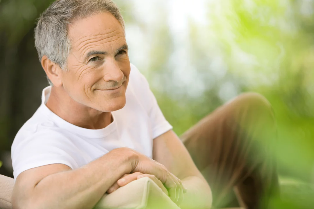 Starší muž v bílém tričku sedí venku a přemýšlí s úsměvem, obklopen zelení. Relaxace a klid na čerstvém vzduchu.