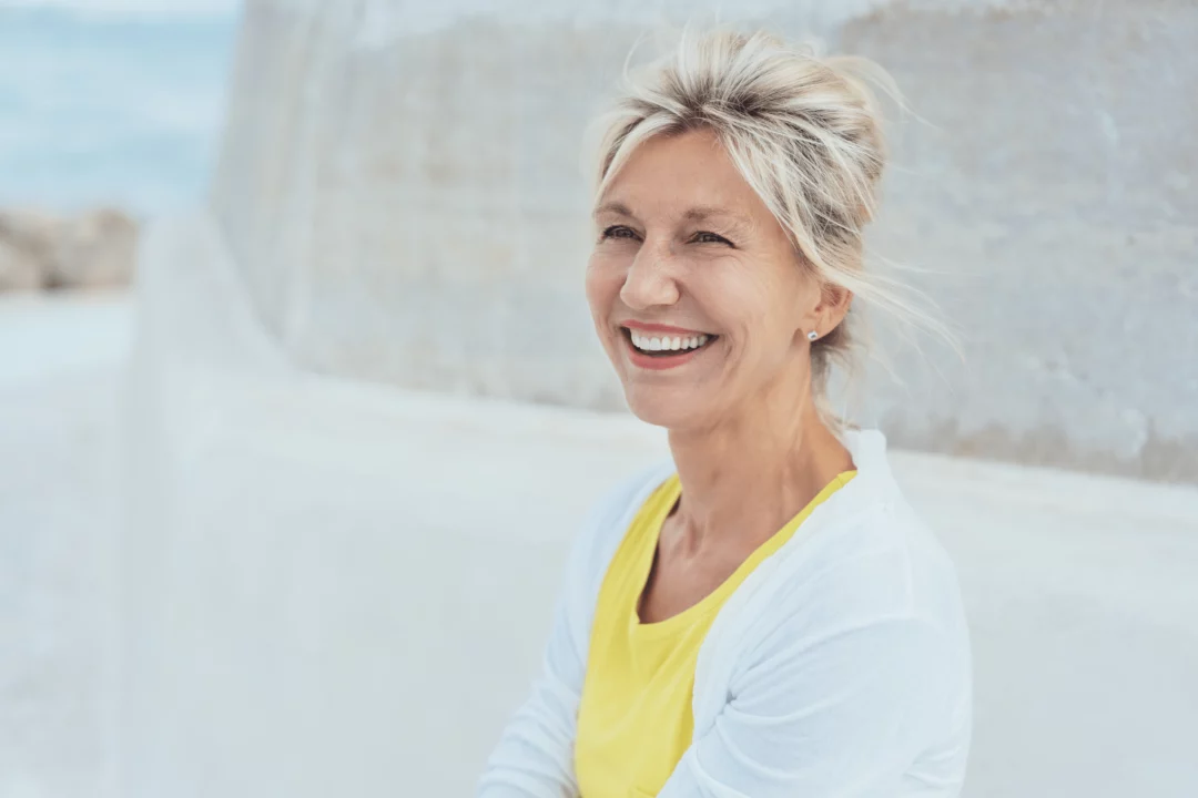 Usměvavá starší žena v žlutém tričku venku, bijící slunce, neformální portrét, modré pozadí, radostný výraz na tváři.