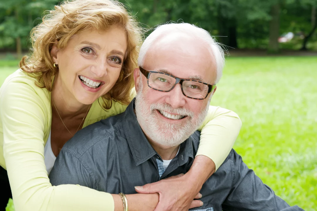 Šťastný seniorský pár v přírodě se usmívá a objímá, vyzařuje radost a spokojenost na slunečném trávníku v parku.