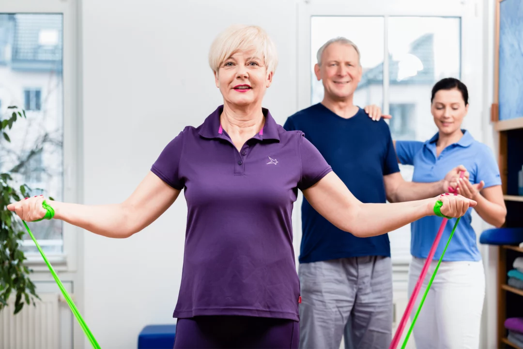 Starší lidé cvičí s odporovými gumami ve skupině v tělocvičně, zaměřují se na zdraví a pohyblivost.