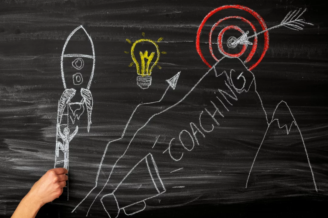 Kresba na tabuli s raketou, žárovkou a horami symbolizujícími coaching a růst k dosažení cíle s terčem a šipkou.