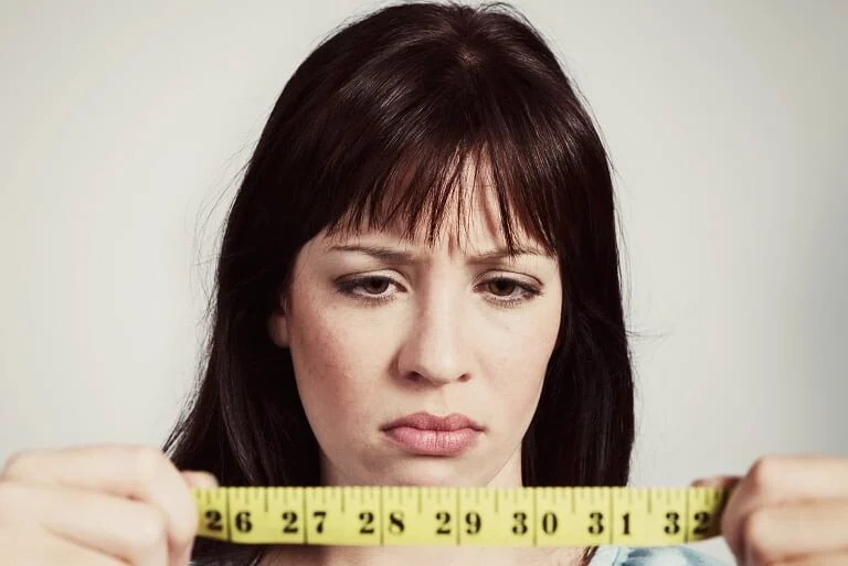 Žena s vážným výrazem drží metr, zamyšleně hledí na čísla. Porovnávání délky, zaměření na přesnost měření.