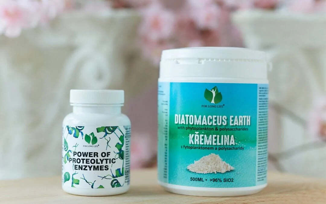 Dvě lahve doplňků stravy: enzymy a křemelina, na dřevěném povrchu s rozmazaným pozadím květů.