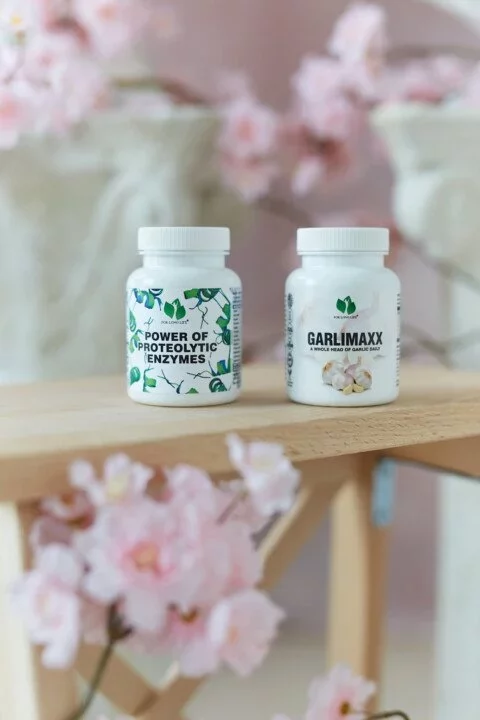 Láhve s doplňky stravy Power of Proteolytic Enzymes a Garlimaxx na dřevěné polici, obklopené růžovými květinami.