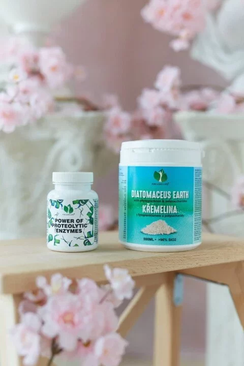 Láhve s proteolytickými enzymy a křemelinou na dřevěném stole s květinovou dekorací v pozadí.