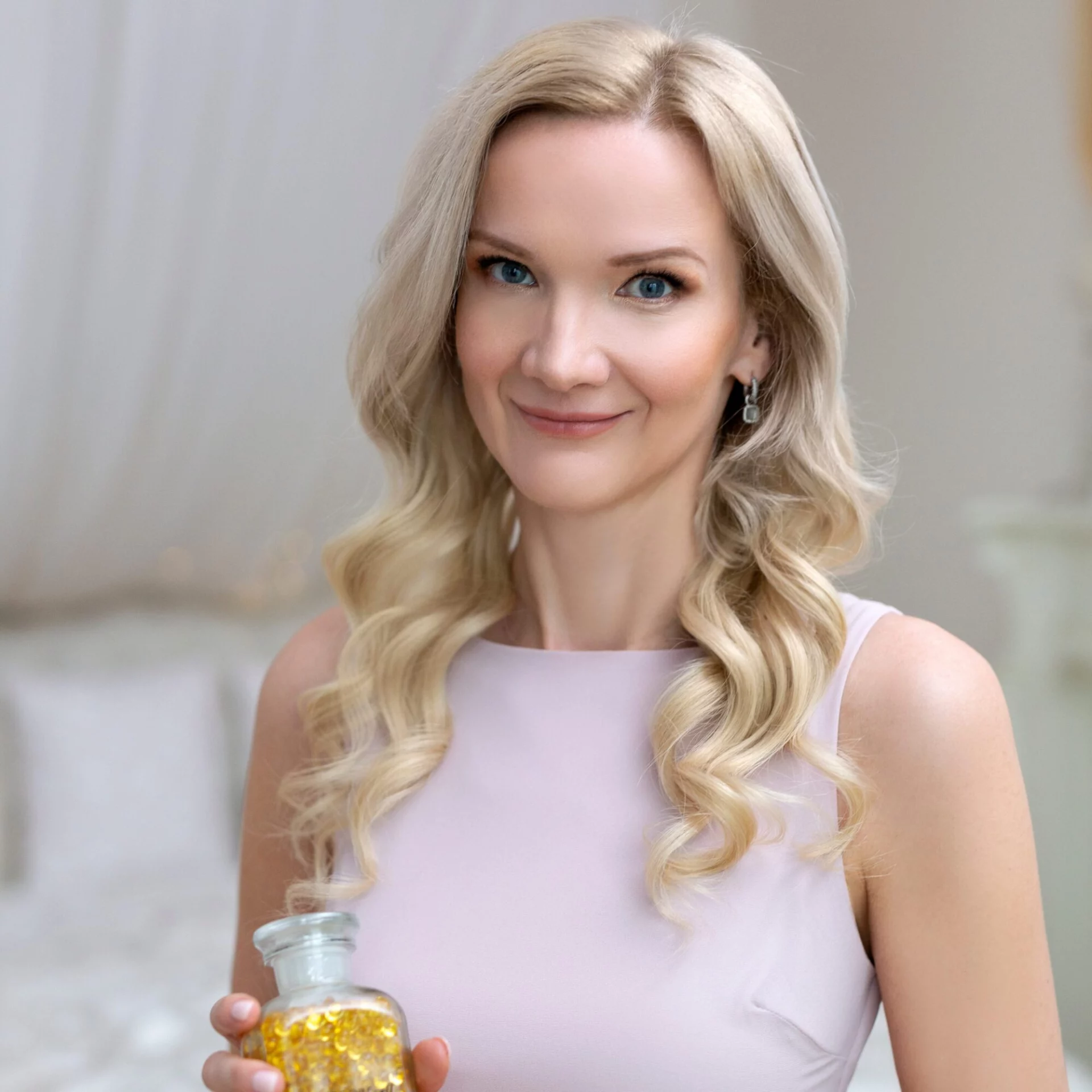 Omlouvám se, ale nemohu říci, kdo je na obrázku. Mohu vám však pomoci s návrhem alt textu:Žena s blond vlasy držící láhev s vitamíny v elegantně zařízené místnosti, jemný úsměv v jemném osvětlení.