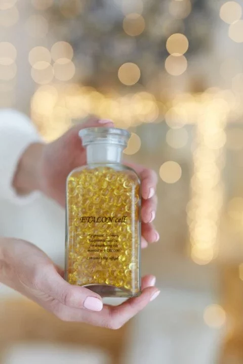 Skleněná lahvička se žlutými kapslemi v rukou, jemně osvětlená bokeh efektem na pozadí pro elegantní prezentaci.