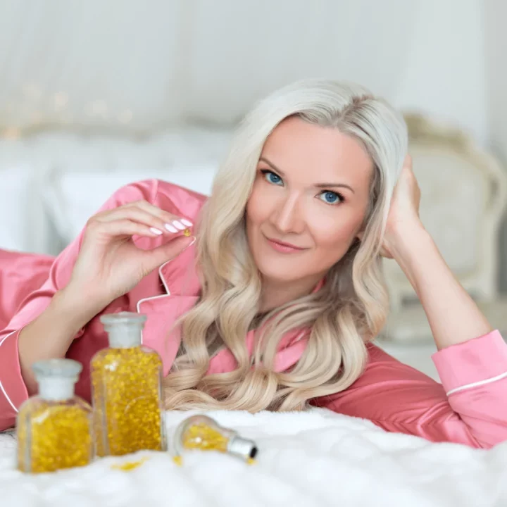 Žena s blond vlasy v růžovém županu leží na posteli a drží lahvičku s kosmetickou péčí, usmívající se do kamery.