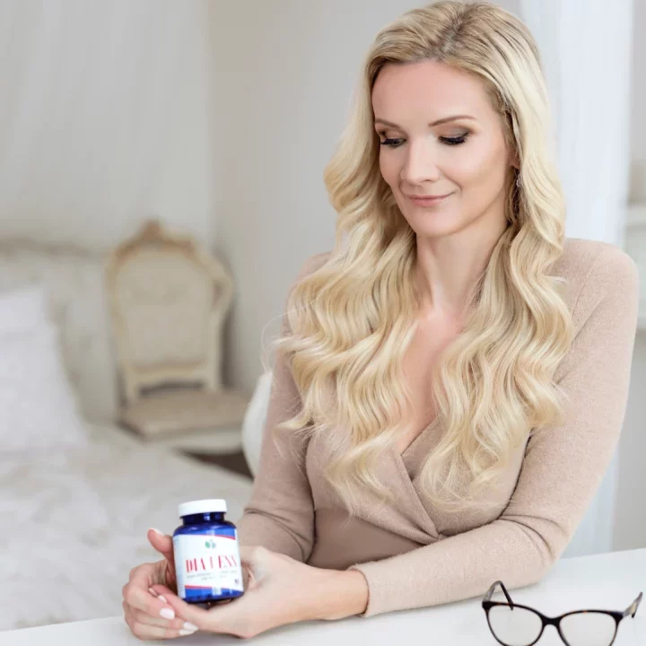 Žena s dlouhými blond vlasy drží lahvičku doplňku na bílé stolku v elegantním, světle dekorovaném interiéru.