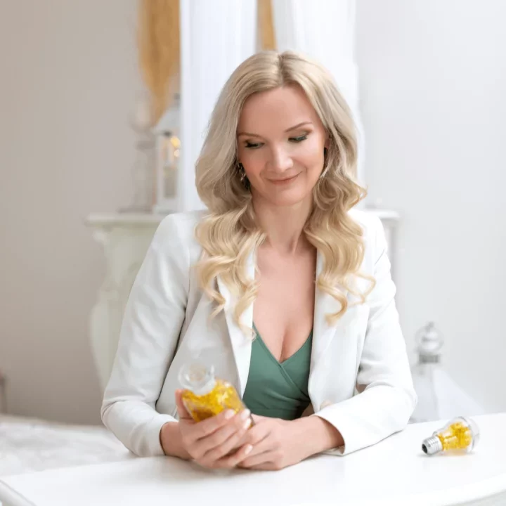 Žena v bílém sáčku se usmívá a drží láhev s olejem na kosmetickém stole, světlý interiér na pozadí.