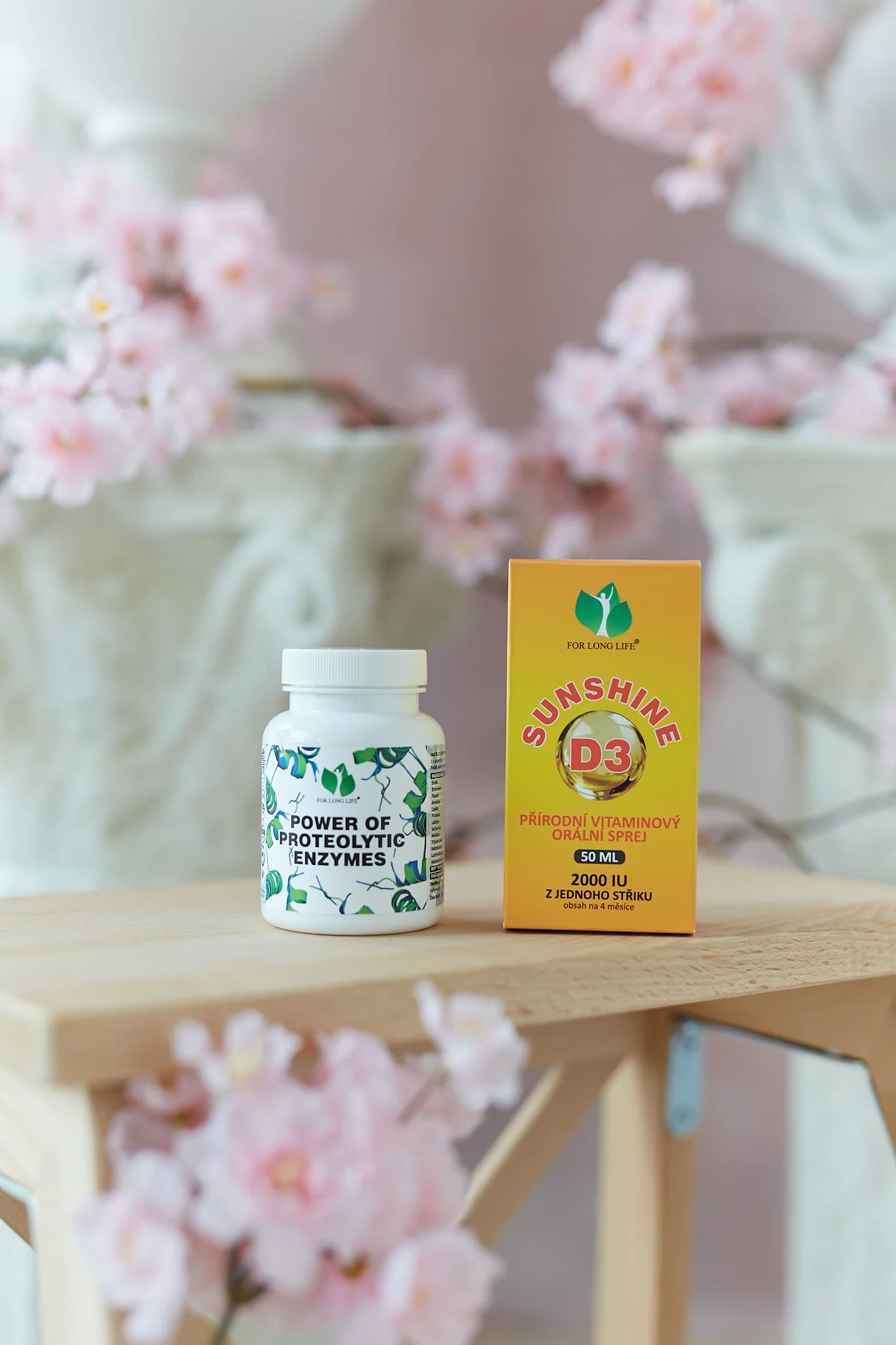 Dva zdravotní produkty na dřevěném povrchu bílá lahvička Power of Proteolytic Enzymesa žlutá krabička spreje s vitamínem D3, obklopená růžovými květy.