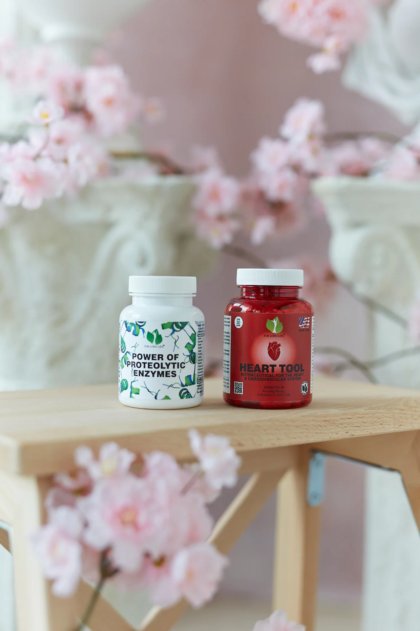 Dvě lahvičky s vitamíny, „Power of Proteolytic Enzymes“ a „Heart Tool“, vystavené na dřevěném stole zdobeném růžovými květy.